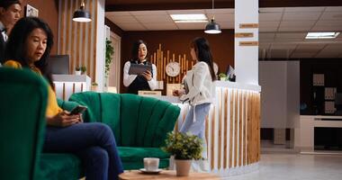 Asian guest signing check in papers on tablet at reception, registering her room confirmation while bellboy is helping with her luggage. Five star hotel staff team friendly welcoming client. photo