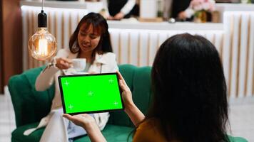 Traveler checking isolated greenscreen template on tablet, sitting with her friend in lounge area and looking at blank copyspace template. Asian guest using isolated mockup. Tripod shot. photo