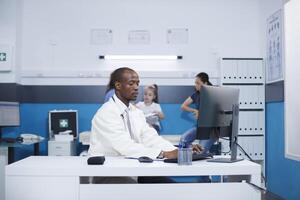 Determined African American doctor writing down his medical information on a desktop computer. A nurse tends to a girl and her mother in a modern examination room in the backdrop. photo