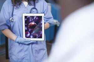 Close-up shot of a caucasian person holding a tablet displaying an image of a virus cell. Selective focus of a bacteria shown on a digital device held by an individual wearing blue scrubs. photo