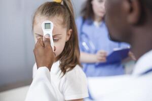 Close-up shot of an African American doctor using a thermometer to check a young girl fever. The youngster is treated in a hospital room after taking a temperature to get a diagnosis. photo