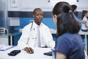 Selective focus on the african american doctor using the computer while speaking with the caucasian female patient in the clinic. The atmosphere is professional and reassuring. photo