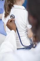 Close-up image of a doctor listening to heartbeat of a child with stethoscope. At doctor appointment in clinic office, an African American physician examines health of a young girl. photo