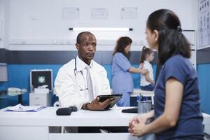 In hospital room, African American male doctor in a lab coat explains healthcare and medicine to a Caucasian woman. A female nurse performs a medical checkup on a small girl in the background. photo