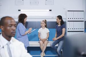 Image shows a mother and daughter seated on a hospital bed, receiving medical consultation from a female nurse practitioner. Communication with patient regarding health. Background focus. photo