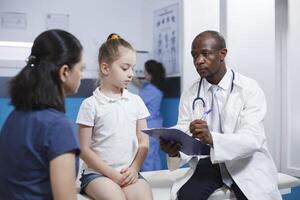 African American doctor recording information on clipboard about small child. In clinic room, little girl and her mother having a healthcare checkup. Medical treatment in a professional context. photo