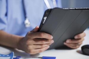 Selective focus of an individual dressed in blue scrubs sitting at a desk, grasping a tablet. Close-up shot of a Caucasian female holding an electronic gadget for medical research. photo