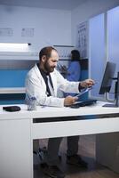 Detailed image of male doctor at the clinic desk going over and analyzing his notes. Caucasian man utilizing desktop computer and clipboard as he prepares for patient medical consultations. photo