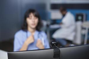 Nurse at hospital is using computer up close to conduct a video chat. In the clinic office, a Caucasian woman is seated and participating in a video conference. Foreground sharp, background fuzzy. photo