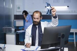 joven médico avaro un radiografía imagen y escritura notas en su bloc. masculino caucásico cuidado de la salud profesional es mostrado en el fotografía examinando un cofre escanear de un paciente. foto