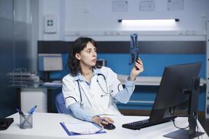 dedicado médico examinando un Connecticut escanear de un paciente, tomando notas en escritorio computadora. el imagen muestra caucásico hembra cuidado de la salud profesional inspeccionando un cofre radiografía imagen de un individual. foto