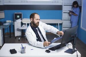 Image shows a committed male doctor reviewing and analyzing his notes at the clinic desk. Young man getting ready for patient medical consultations while using a desktop computer and clipboard. photo