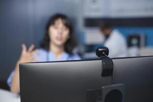 Selective focus on computer being used by nurse practitioner at a hospital for a video call. Caucasian woman is seated in the clinic office, participating in a virtual conference. Close-up shot. photo