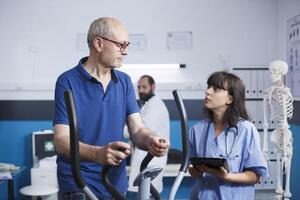 Physiotherapy being administered to patient as he pedals on stationary bike, with assistance of female nurse dressed in scrubs. Healthcare assistant holds tablet to relay medical results to old man. photo