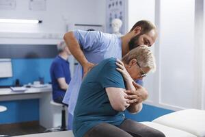 Osteopathic doctor cracking back and shoulder bones for retired old woman at chiropractic consultation. Chiropractor giving assistance to elderly patient for recovery and rehabilitation. photo