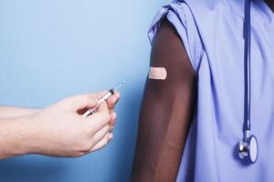 Close-up of caucasian hands with a syringe and needle vaccinating a black person. Doctor administers vaccination shot to African American nurse, who is wearing an adhesive bandage. photo