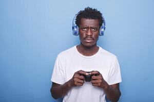 retrato de africano americano chico jugando vídeo juegos con controlador mientras vistiendo auriculares en estudio. negro hombre escuchando a música y participación palanca de mando para juego jugar en consola, curioso a cámara. foto