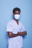 Nurse with crossed arms wearing face mask and gloves, looking at camera in studio. Man working as specialist standing over blue background, having protection against covid 19 pandemic. photo