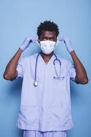 Portrait of a medical specialist putting on face mask to defend against the covid 19 epidemic. Man with blue scrubs, stethoscope, and gloves staring at camera, working as a nurse. photo