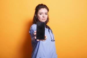 Caucasian woman in nurse uniform is holding smartphone with blank display. Medical assistant stands in front of isolated backdrop and looks at camera while showing empty screen of mobile device. photo