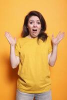 Frightened brunette lady showing face expression of despair, standing over orange background. Shocked woman feeling scared and afraid with hands up while looking at camera. photo
