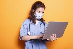 Healthcare specialist wearing face mask looking at laptop and wearing uniform and stethoscope. Medical assistant using personal computer during pandemic while standing over isolated background. photo