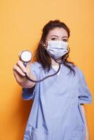 Close-up of female physician wearing blue scrubs grasping stethoscope in front of isolated background. Nurse with face mask gripping a medical instrument towards camera in studio. photo