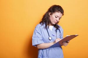 Doctor in blue scrubs writing on clipboard papers, standing against isolated orange background. Female nurse with stethoscope signing documents on medical insurance for patients. photo