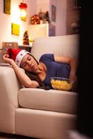 Woman with santa hat sleeping on couch after watching winter entertainment movie on television in xmas decorated kitchen. Caucasian female celebrating december season enjoying christmas holiday photo