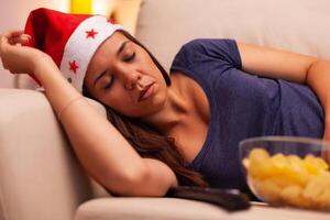 Adult person with santa hat sleeping on sofa after watching december entertainment movie on television in xmas decorated kitchen. Woman celebrating christmas holiday enjoying winter season photo