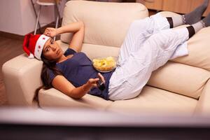 Relaxed woman resting on couch watching christmas movie series on television celebrating christmastime in x-mas decorated kitchen. Girl wearing santa hat enjoying winter season at home photo