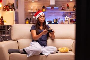 mujer acostado en sofá jugando en línea videojuego utilizando juego de azar palanca de mando durante virtual juego competencia disfrutando invierno fiesta en Navidad decorado cocina. niña celebrando Navidad temporada foto