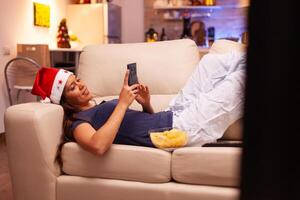 niña mensajería con amigo utilizando teléfono inteligente descansando en sofá en Navidad decorado cocina. mujer hojeada en social medios de comunicación, mensajes de texto durante Navidad fiesta disfrutando invierno temporada foto