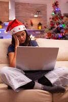 Tired exhausted woman falling asleep on couch in xmas decorated kitchen while reading business email on computer. Caucasian female celebrating christmas holiday enjoying winter season photo