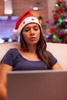 Girl looking at laptop screen reading business email during christmas holiday sitting comfortable on sofa in xmas decorated kitchen. Woman adult browsing on social media. Winter season photo