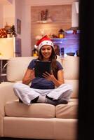 Adult woman wearing santa hat sitting in lotus position on couch while browsing on social media using tablet during christmastime in xmas decorated kitchen. Caucasian female enjoying christmas holiday photo