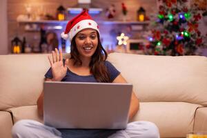 Girl greeting remote friend during online videocall conference meeting on laptop resting on couch in xmas decorated kitchen. Woman celebrating christmas holiday enjoying winter season photo