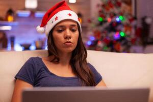 Woman looking at laptop screen reading business email during christmas holiday resting comfortable on couch in xmas decorated kitchen. Caucasian female browsing on social media. Winter season photo