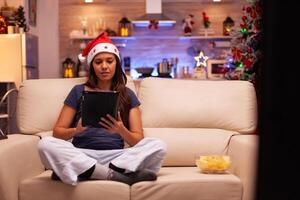 Adult woman wearing santa hat sitting in lotus position while browsing on social media using tablet during christmastime in xmas decorated kitchen. Caucasian female celebrating christmas holiday photo