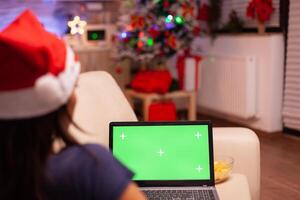 mujer sentado en sofá en Navidad decorado cocina participación verde pantalla burlarse de arriba croma llave ordenador portátil computadora con aislado mostrar. adulto persona disfrutando Navidad fiesta celebrando invierno temporada foto