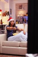 Girl wearing red santa hat holding remote searching funny xmas comedy on television during christmastime enjoying winter season on x-mas decorated kitchen. Woman adult celebrating christmas holiday photo