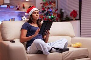 Woman sitting in lotus position on couch looking on social media writing xmas email messaging with friends using tablet computer. Adult person celebrating christmas season enjoying winter holiday photo