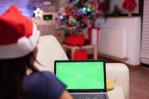 Caucasian female looking at mock up green screen chroma key laptop with isolated display resting on couch in xmas decorated kitchen. Woman enjoying winter season celebrating christmas holiday photo