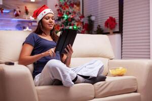 Woman sitting in lotus position on sofa browsing on social media writing xmas email messaging with friends using tablet computer. Adult person celebrating christmas season enjoying winter holiday photo