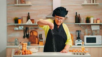 Woman is sifting flour on wooden table in modern kitchen. Happy elderly baker with bonete preparing raw ingredients to baking homemade cake sprinkling, sieving wheat flour by hand photo