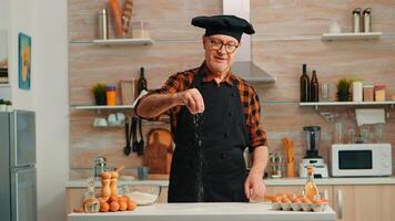 Senior baker in home kitchen putting wheat flour over wooden table. Retired elderly chef with bonete and apron sprinkling, sieving sifting raw ingredients by hand baking homemade pizza, bread. photo