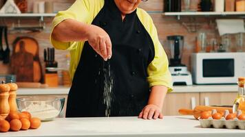 Old lady preparing food in home kitchen spreading flour for baking a recipe. Retired elderly chef with uniform sprinkling sieving sifting ingredients by hand on table cooking homemade pizza bread photo