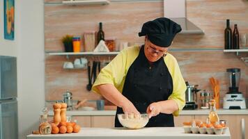 Mature woman baker mixing by hand cracked eggs with flour in home kitchen following traditional recipe. Retired elderly chef with bonete kneading in glass bowl pastry ingredients baking homemade cake photo