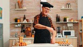 mayor panadero enharinado mesa a hornear y hacer delicioso galletas. retirado mayor cocinero con bonete y delantal en uniforme aspersión tamizado cernido crudo ingredientes por mano horneando hecho en casa pizza, un pan foto