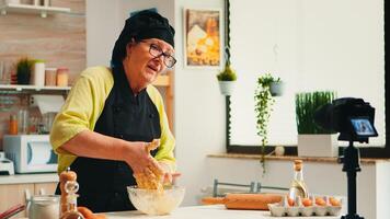 Mature experienced baker recording culinary recipe tutorial using modern camera in kitchen wearing bonete and apron. Influencer chef using internet technology communicating on social media. photo